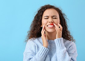 Woman with curly brown hair in blue sweater closing her eyes and touching her inflamed gums in pain